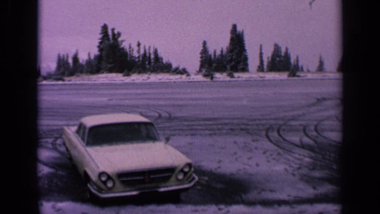 A white mid-century car appears at the edge of a frozen Washington roadway, with circular tire tracks and dark evergreen trees behind it.