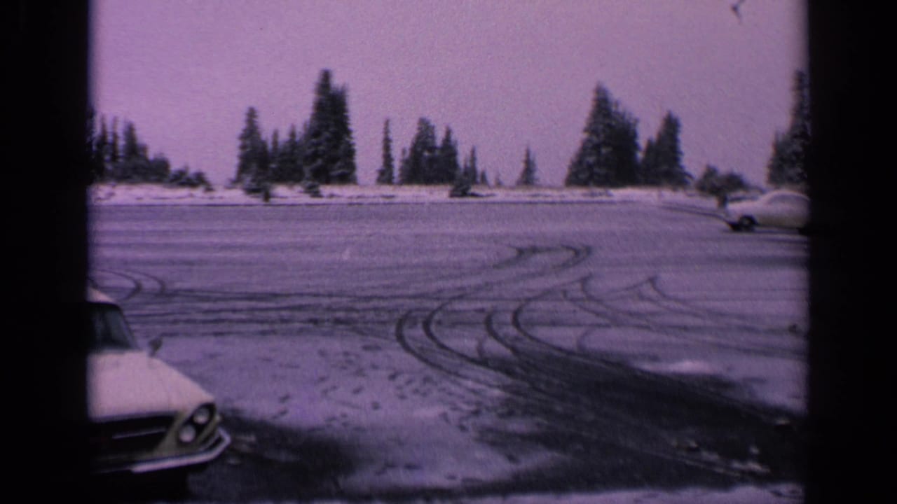 Snowy parking lot with curved tire tracks, evergreens, and the front of a car at the left edge.