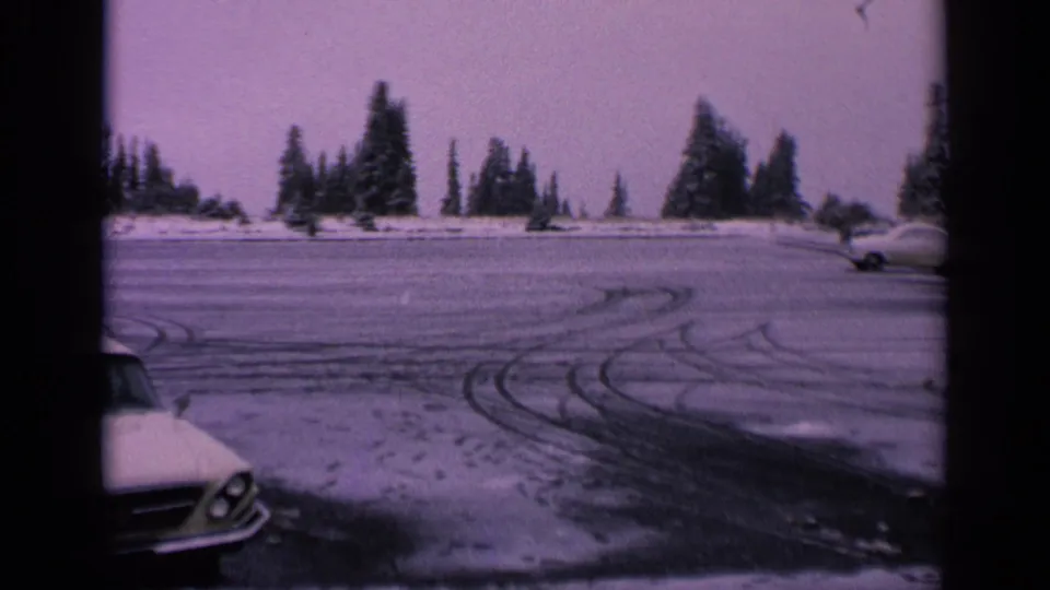 1963 Washington 8mm frame showing a snowy parking lot, tire tracks, evergreen trees, and a partial classic car at the left edge.