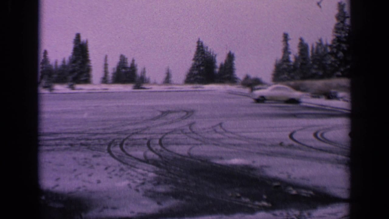 1963 Washington 8mm frame of a pale classic car near the edge of a snowy parking lot lined with evergreen trees.