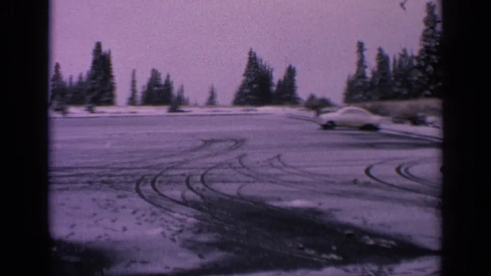 1963 Washington 8mm frame of a pale classic car near the edge of a snowy parking lot lined with evergreen trees.