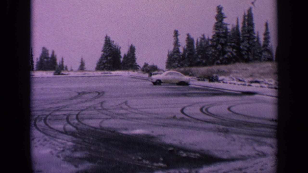 1963 Washington 8mm frame showing tire tracks across a snowy lot and a pale car beside evergreen trees.