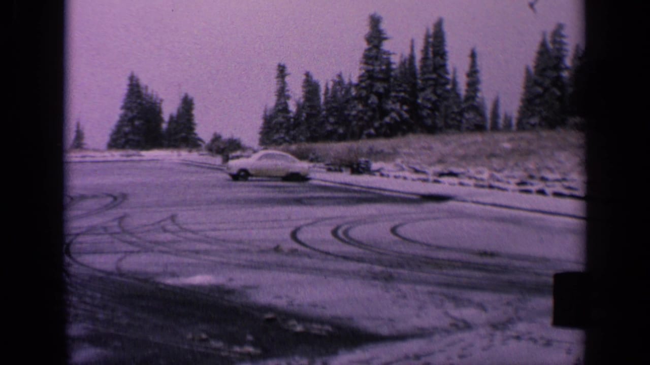 1963 Washington 8mm frame of a pale classic car crossing a snowy parking lot marked by curved tire tracks.