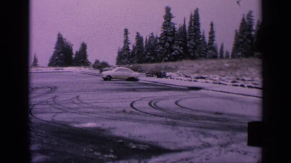 1963 Washington 8mm frame of a pale classic car crossing a snowy parking lot marked by curved tire tracks.