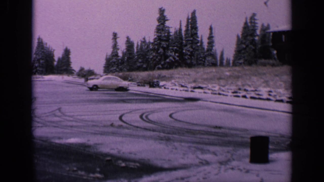 White car on a snowy parking lot with curved tire marks and evergreen trees behind it.