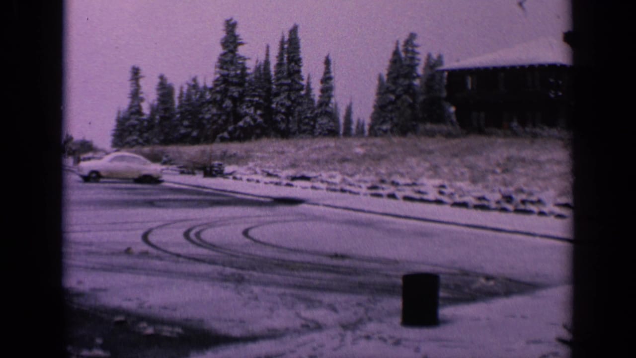 1963 Washington home-movie frame with a white car on icy pavement, visible skid arcs, snow-covered trees, and a hillside structure.