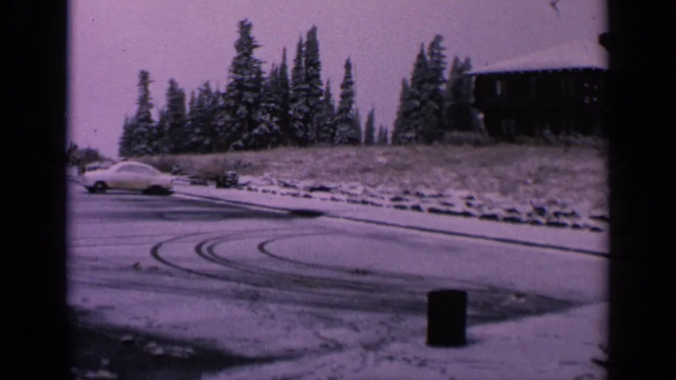 1963 Washington home-movie frame with a white car on icy pavement, visible skid arcs, snow-covered trees, and a hillside structure.
