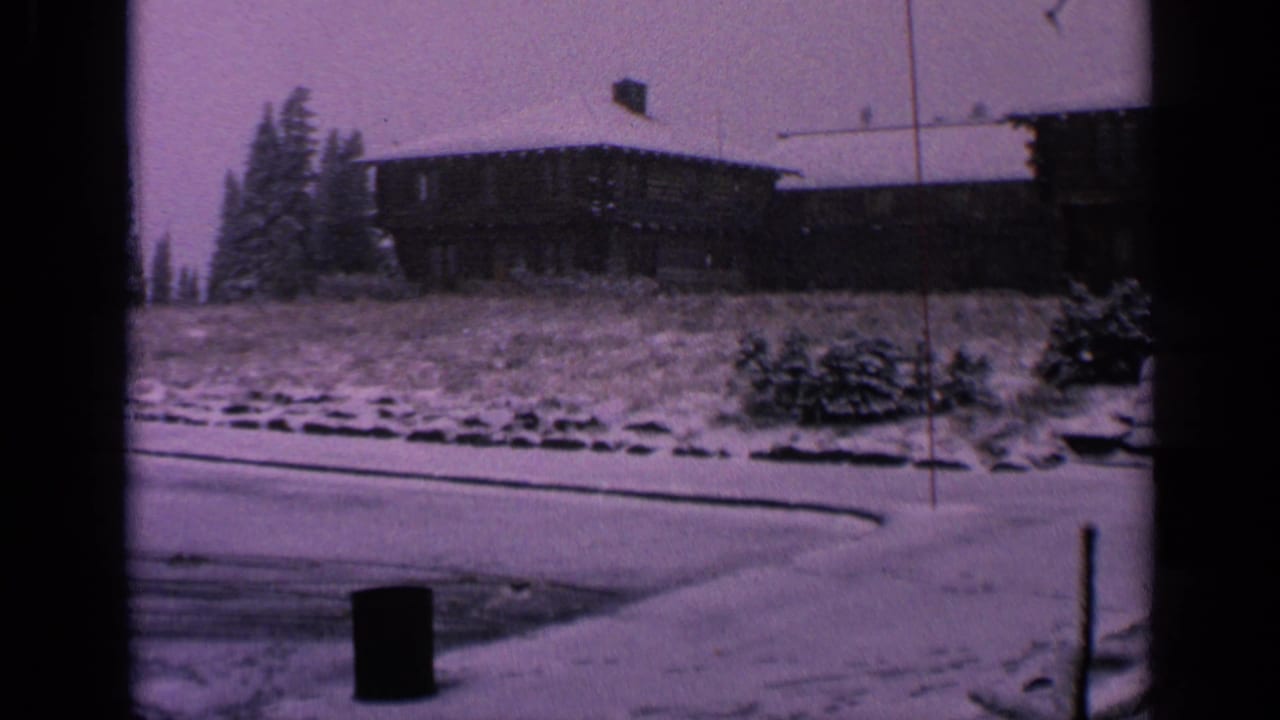 1963 Washington home-movie frame showing a snow-covered parking area, dark tire tracks, evergreens, a hillside building, and roadside posts.
