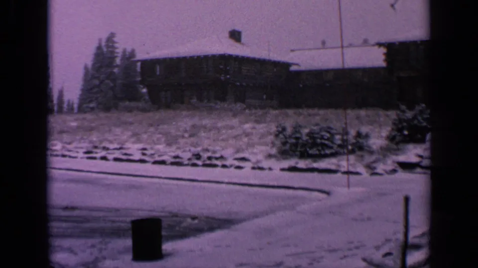 1963 Washington home-movie frame showing a snow-covered parking area, dark tire tracks, evergreens, a hillside building, and roadside posts.