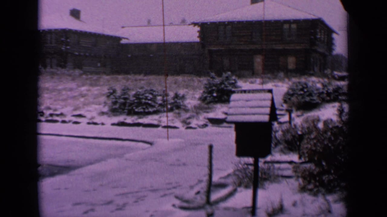 1963 Washington home-movie view of snow-covered log-style homes, roadside shrubs, a small dark structure, and a curved road edge.