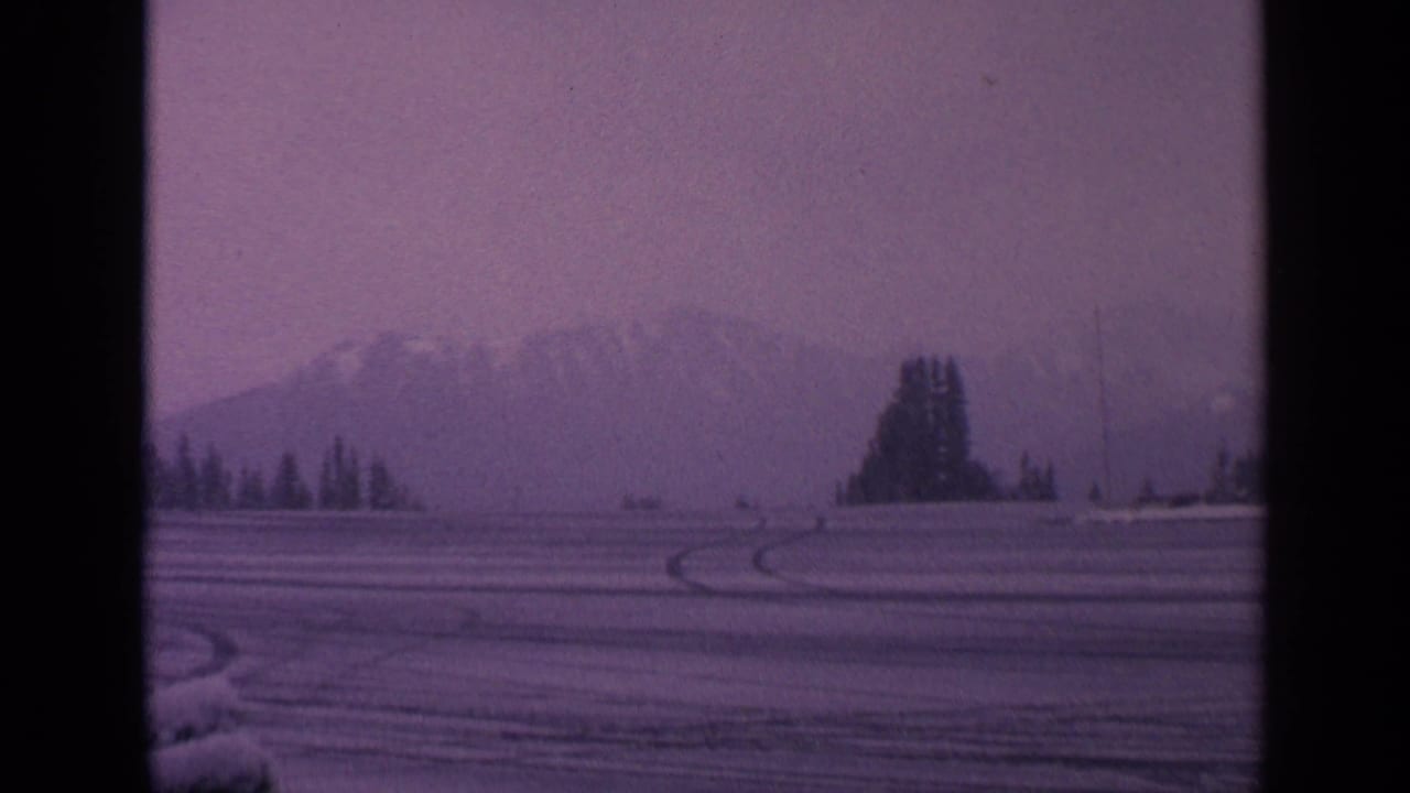 1963 Washington winter landscape with curved vehicle tracks in snow, a tree cluster, and mountains under a hazy sky.