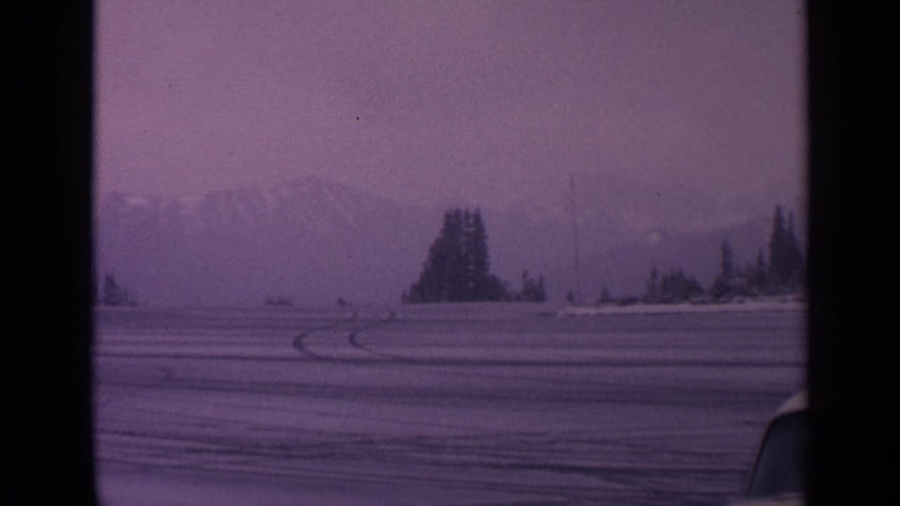 Snowy field with curved tire tracks, evergreen trees, distant mountains, and a partial car roofline.