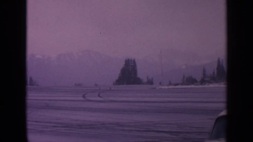1963 Washington 8mm frame showing a snowy open field, curved tire tracks, evergreens, hazy mountains, and a car edge in the foreground.