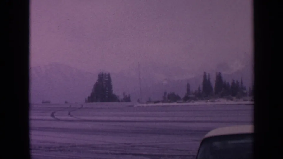 1963 Washington winter landscape frame with a snowy field, evergreen clusters, faint mountains, tire tracks, and a vehicle roofline at lower right.