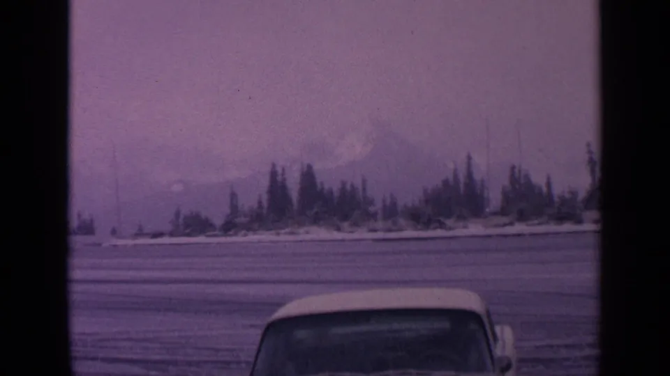 1963 Washington winter frame with an open snowy field, evergreen trees, hazy mountain backdrop, and the roof of a vehicle in the foreground.