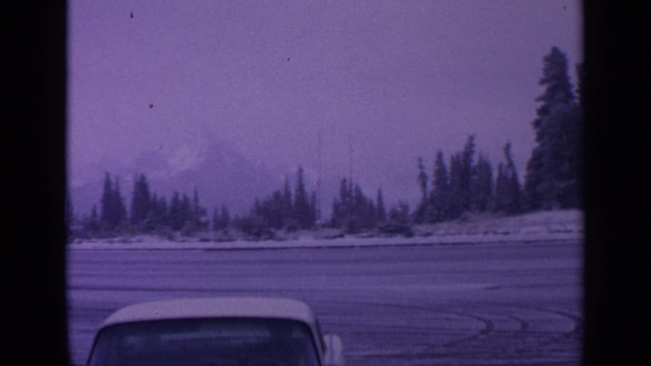1963 Washington winter landscape with evergreen shoreline, faint snow-capped mountain, icy foreground, and partial vehicle roof visible in 8mm frame.