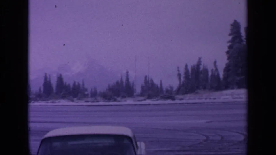 1963 Washington winter landscape with evergreen shoreline, faint snow-capped mountain, icy foreground, and partial vehicle roof visible in 8mm frame.