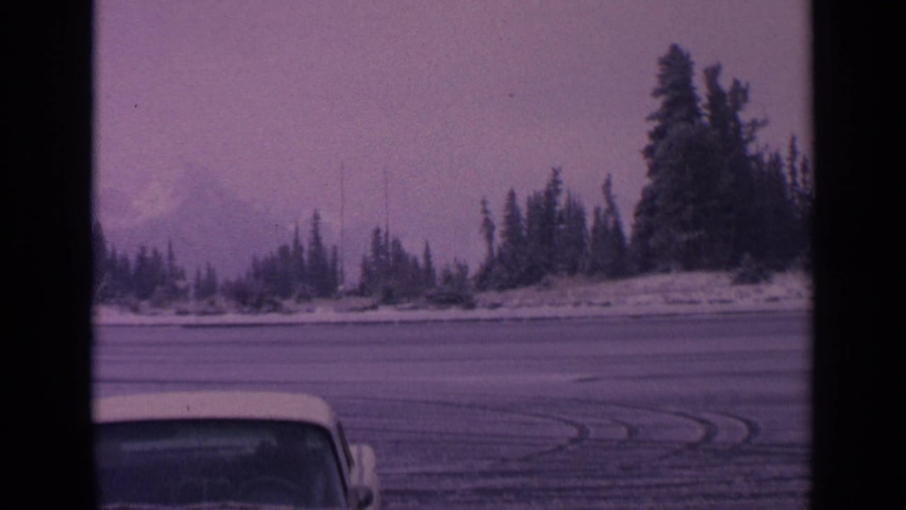 1963 Washington 8mm frame showing curved tire tracks on ice or packed snow, evergreen shoreline, hazy mountain, and partial vehicle foreground.