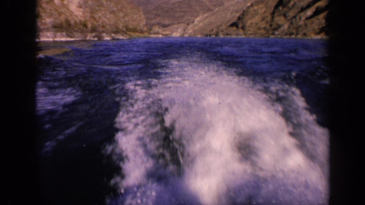 Vintage 8mm Washington river frame showing a broad white boat wake and rocky banks from a moving home-movie viewpoint.