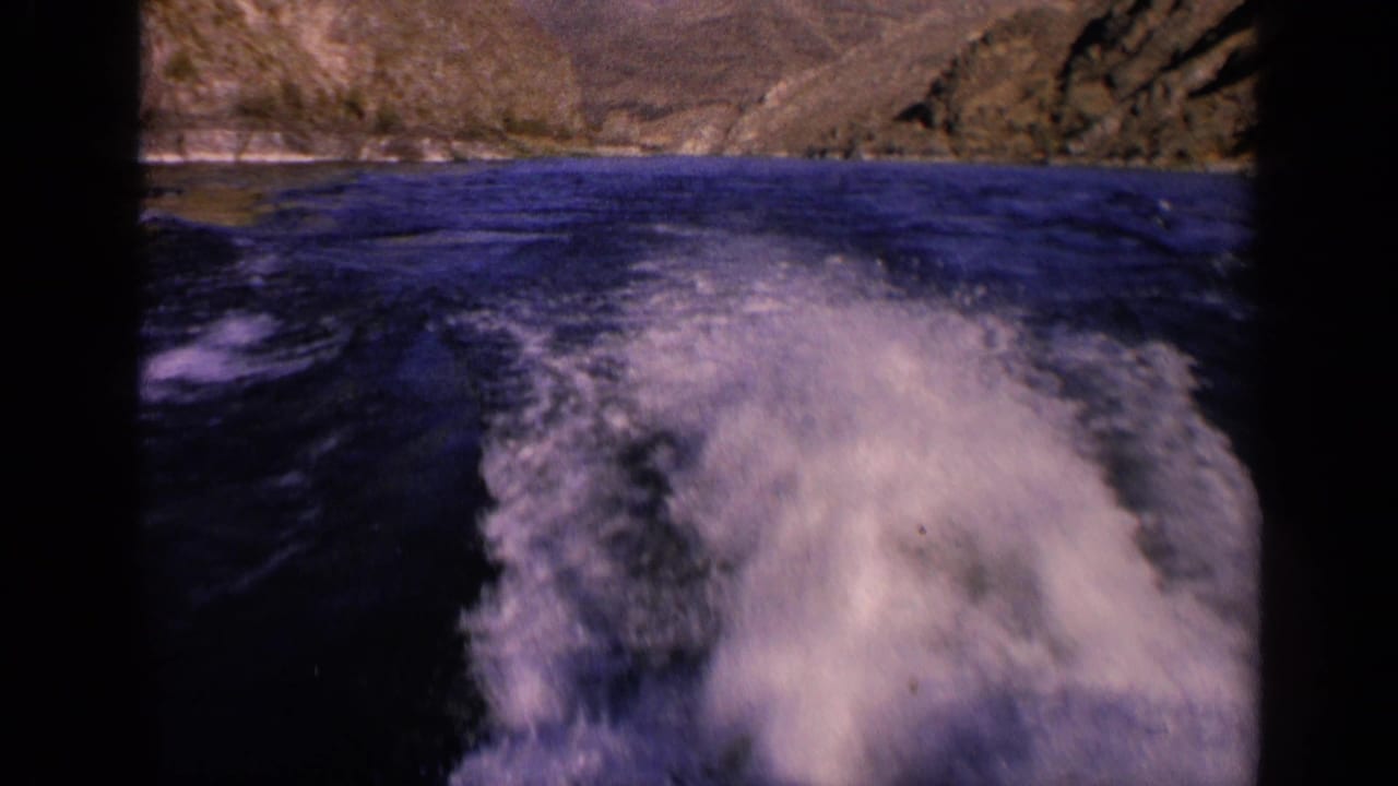 1963 Washington 8mm frame showing a stern view of foamy boat wake trailing across dark water toward rugged shoreline hills.