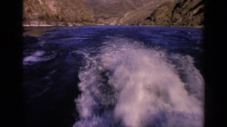 1963 Washington 8mm frame showing a stern view of foamy boat wake trailing across dark water toward rugged shoreline hills.