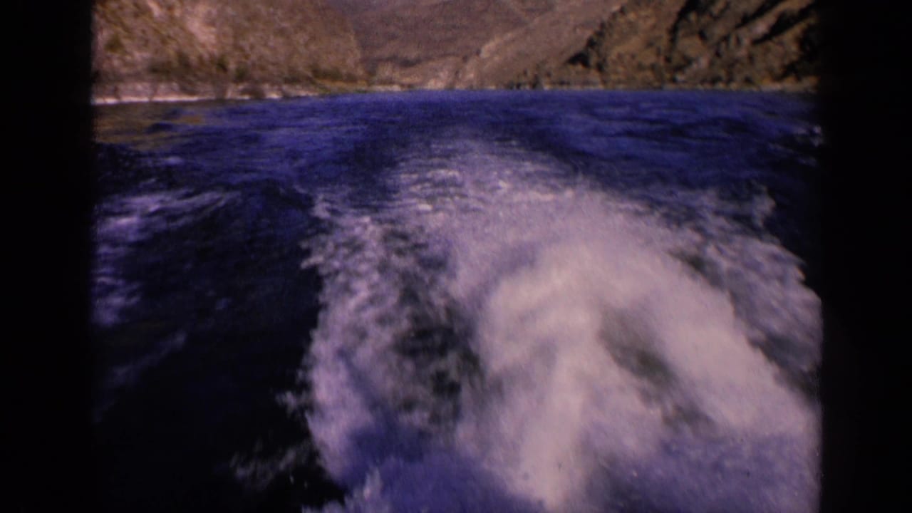 White foam spreads behind a moving boat with rocky shoreline in the background.