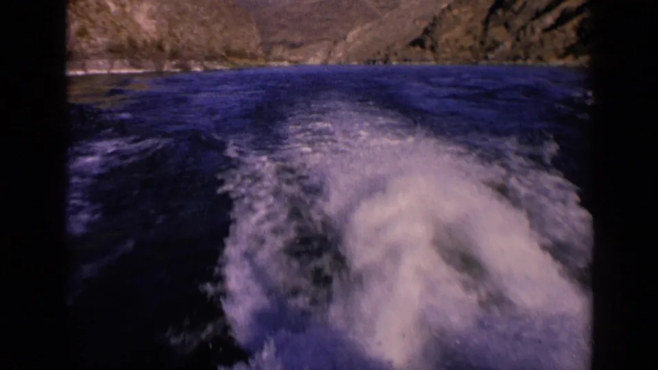 1963 Washington home-movie frame of turbulent white wake spreading over blue water beneath distant rocky hills.