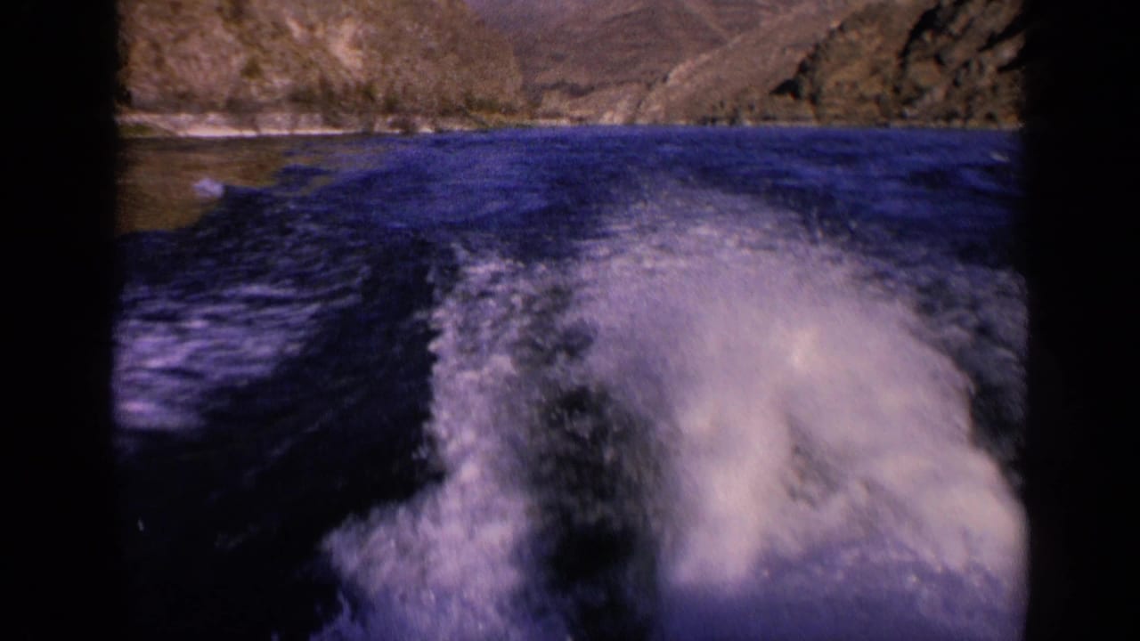 1963 Washington home-movie frame showing a curved boat wake over deep blue water with dry rocky hills in the distance.