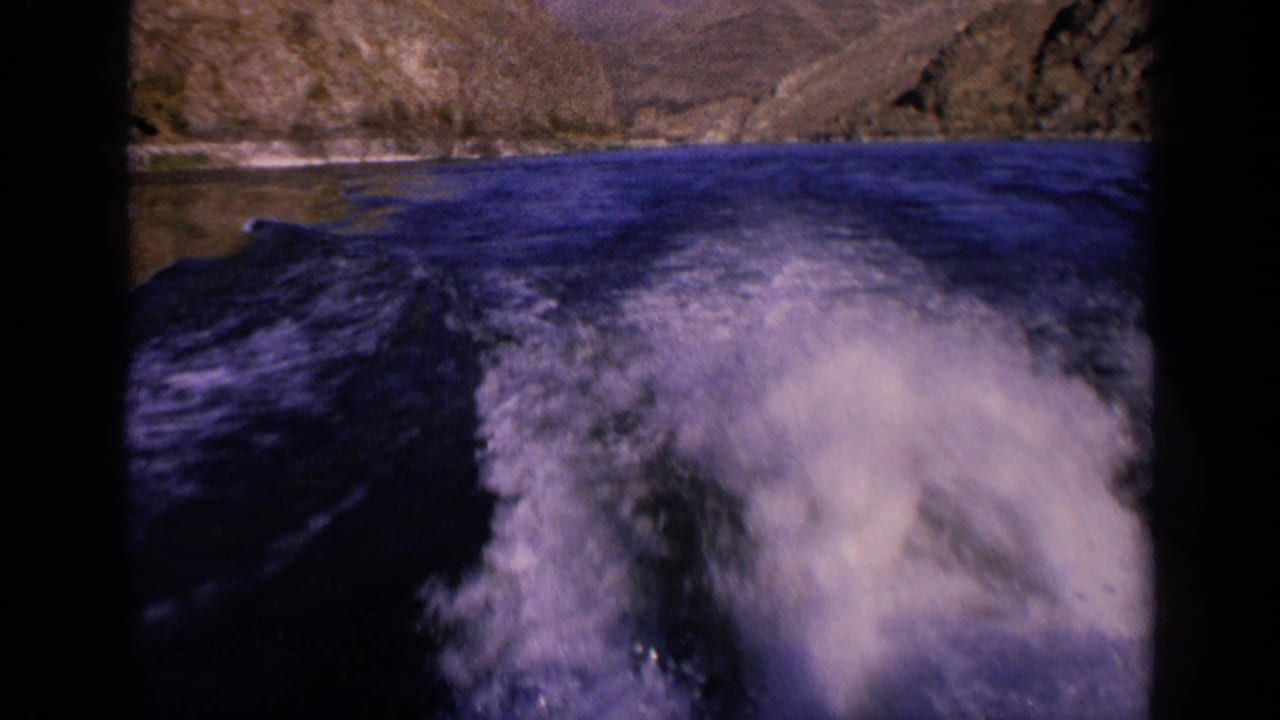 1963 Washington 8mm frame showing a rear view of white boat wake spreading over blue river water below rocky hills.