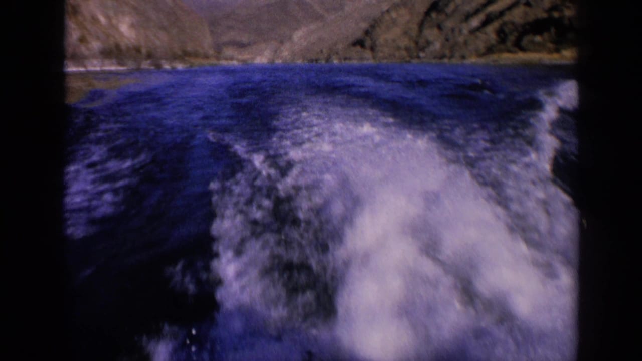 1963 Washington 8mm frame showing a broad foamy wake on blue river water with rocky shore terrain in the distance.