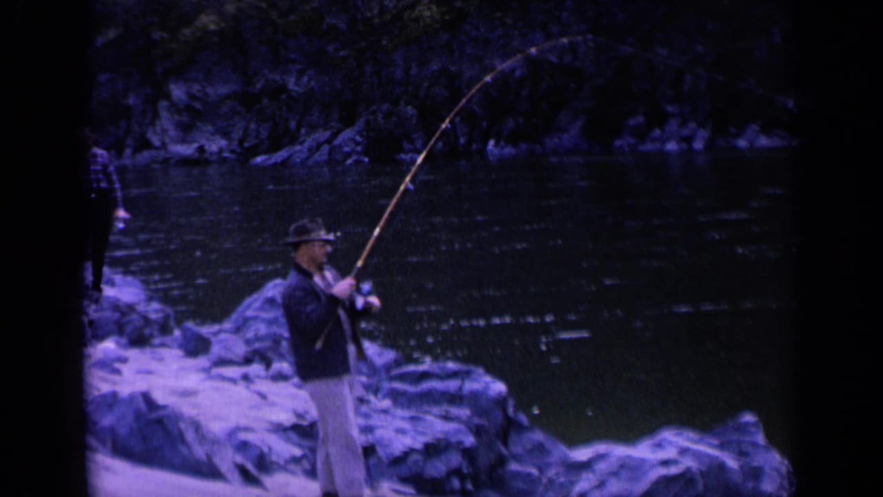 In 1963 Washington home-movie footage, a man in a fedora fishes from a rocky riverbank while another person stands farther back.