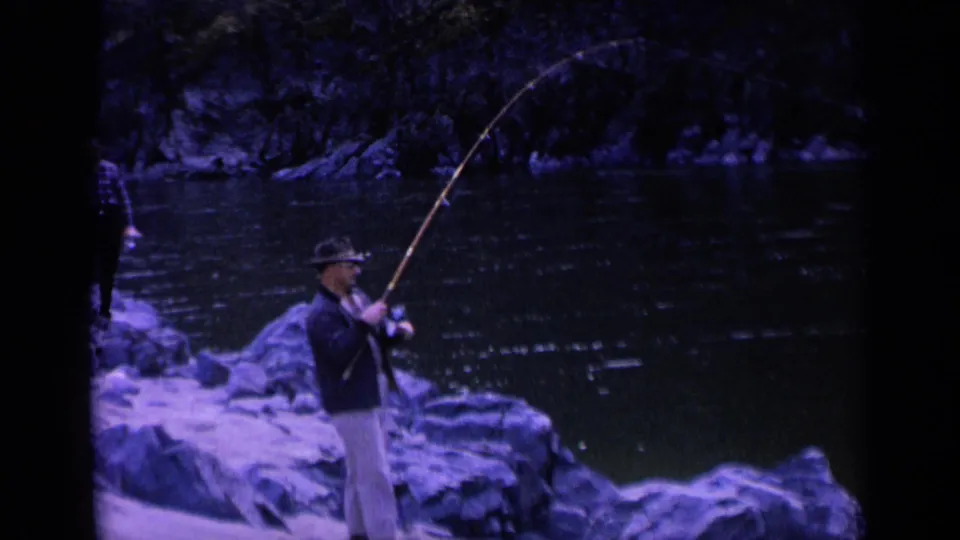 In 1963 Washington home-movie footage, a man in a fedora fishes from a rocky riverbank while another person stands farther back.