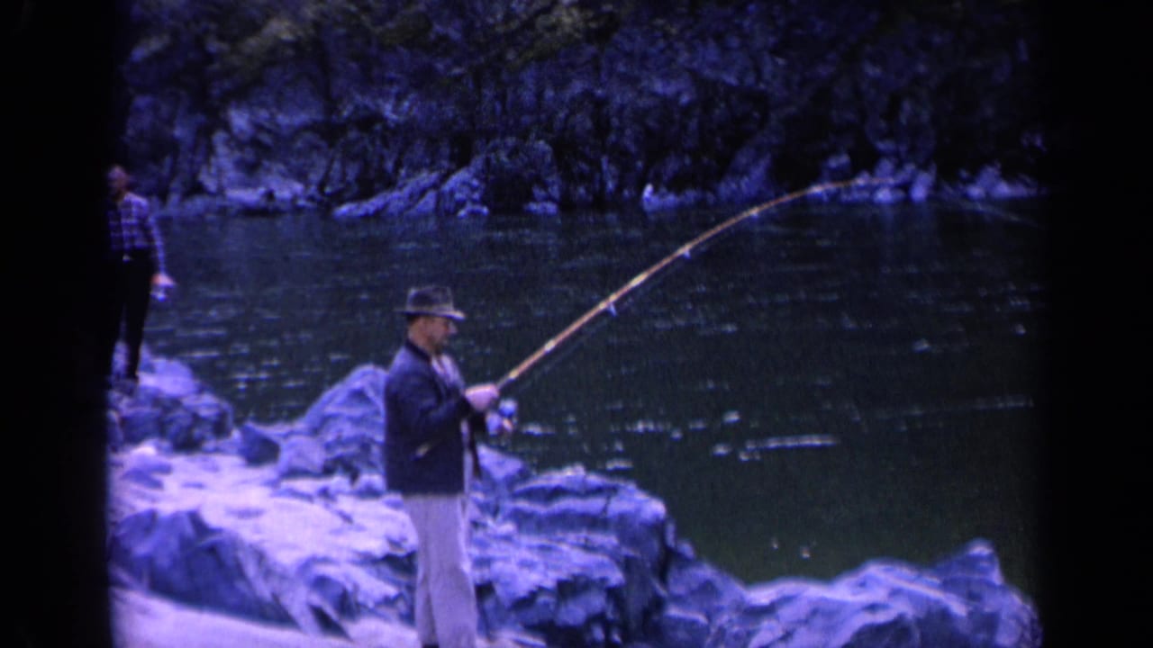 A man in jacket and hat works a long fishing pole beside a Washington river, with a plaid-shirt observer partly visible nearby.