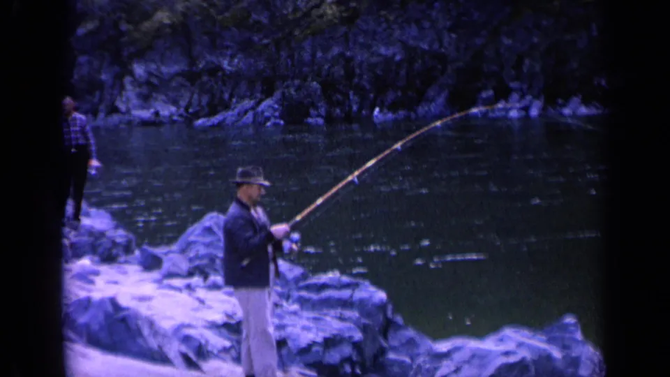 A man in jacket and hat works a long fishing pole beside a Washington river, with a plaid-shirt observer partly visible nearby.