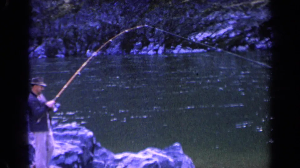 The fisherman remains partly framed at the lower left as his long pole arcs across the river in blue-purple 8mm footage.