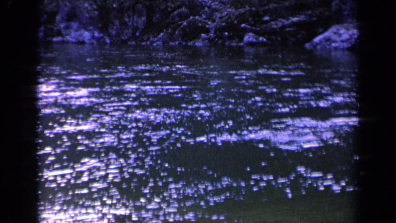 1963 Washington 8mm frame showing churning dark water, bright surface flecks, rocks, and shaded vegetation at the bank.