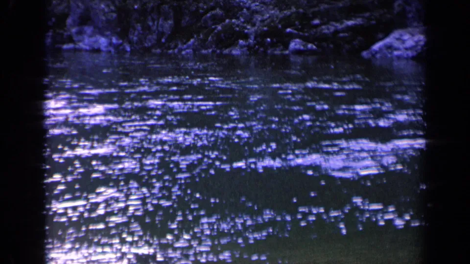 1963 Washington 8mm frame showing churning dark water, bright surface flecks, rocks, and shaded vegetation at the bank.