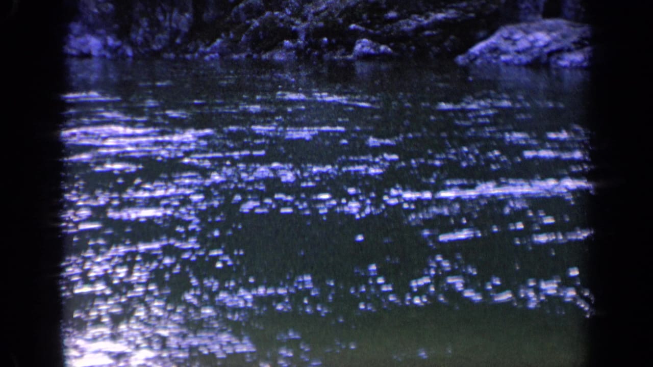 1963 Washington 8mm frame showing bright reflections scattered over dark pond water, with a shaded rocky or leafy bank along the upper edge.