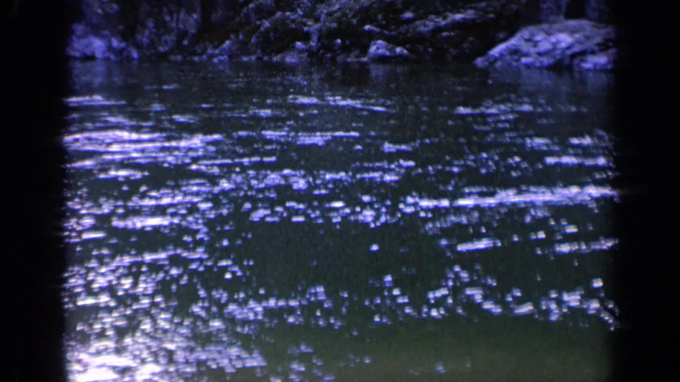 1963 Washington 8mm frame showing bright reflections scattered over dark pond water, with a shaded rocky or leafy bank along the upper edge.