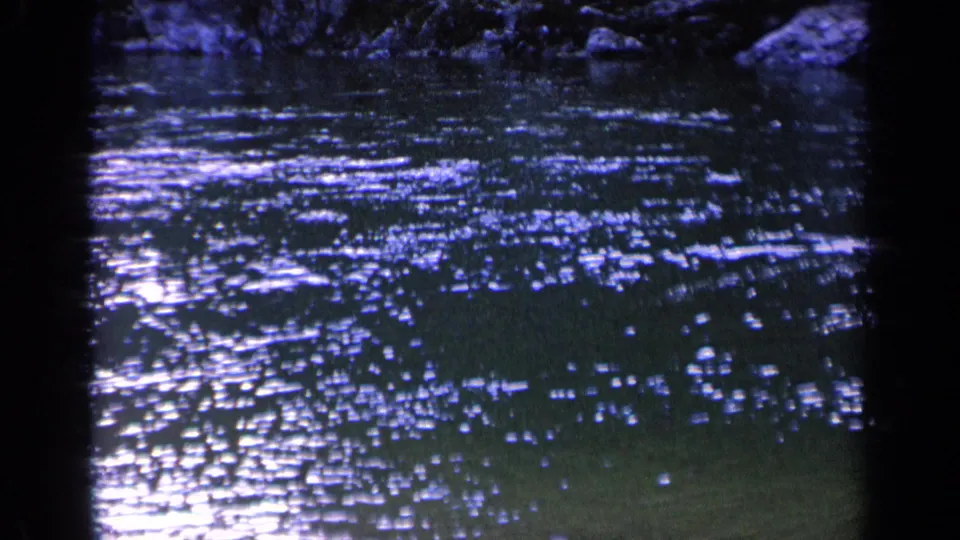 1963 Washington home-movie frame with shimmering reflected light on rippled pond water and a dark natural edge at the top of frame.