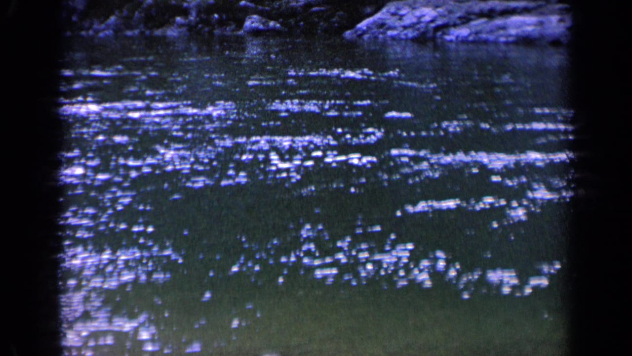 1963 Washington 8mm still showing scattered sun glints on dark pond water, with a shaded bank and rocks visible along the upper margin.