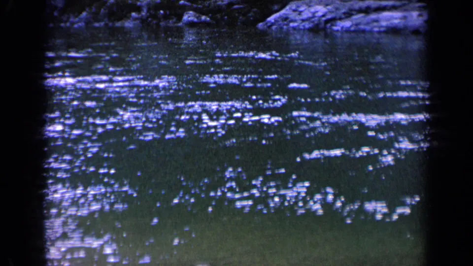 1963 Washington 8mm still showing scattered sun glints on dark pond water, with a shaded bank and rocks visible along the upper margin.