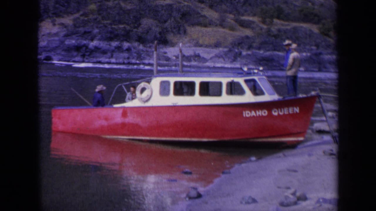 Side view of the IDAHO QUEEN boat with men aboard near a rocky bank.