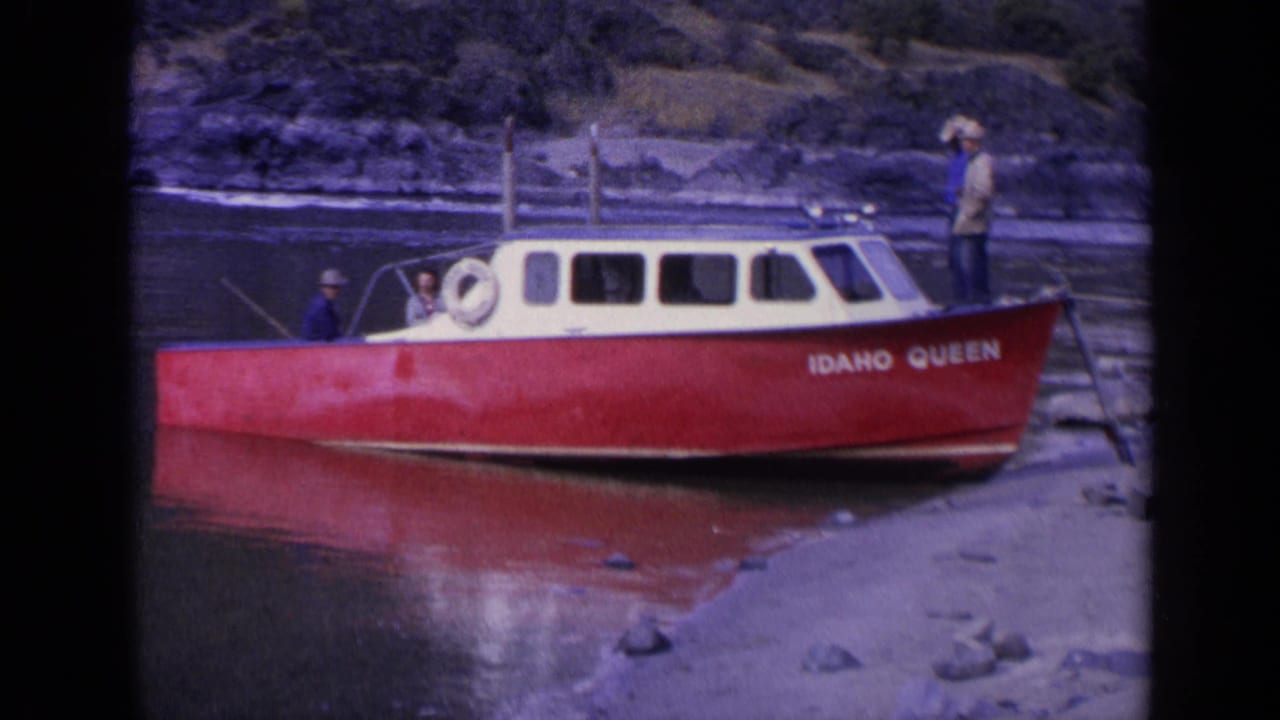 1963 Washington 8mm frame showing the red Idaho Queen boat at shore, with men in hats around the cabin and dark water behind.