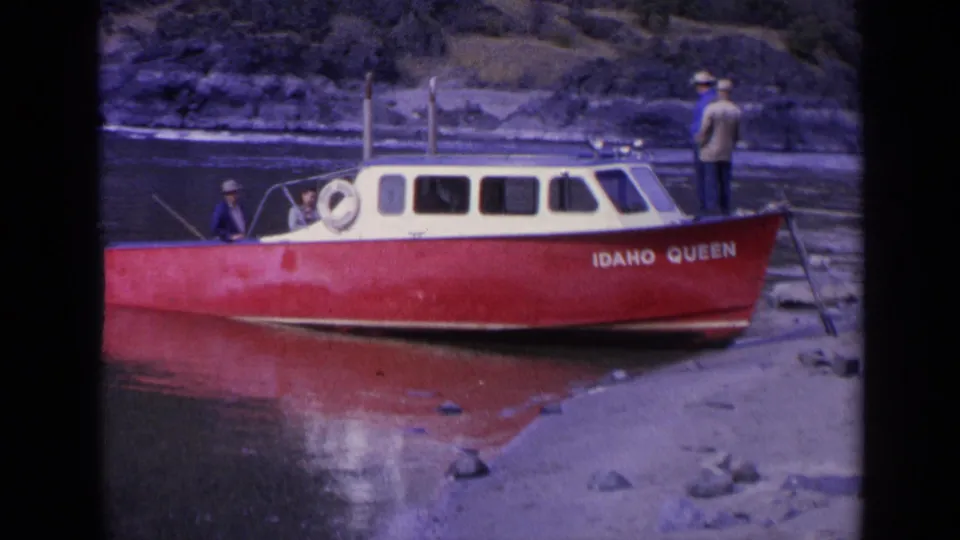 The Idaho Queen sits at a rocky Washington shoreline, its red hull and white cabin visible as several men stand or sit near the deck.