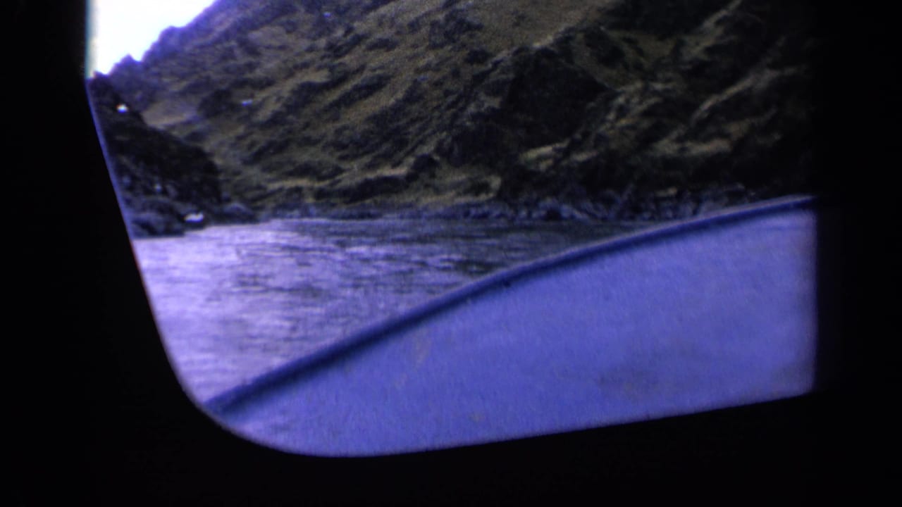 Vessel edge in foreground with river, rocky bank, and tall grassy hills beyond.