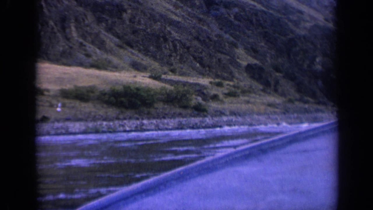 1963 Washington 8mm roadside view showing a river with small white caps, rocky bank, bushes, and a steep mountain slope.
