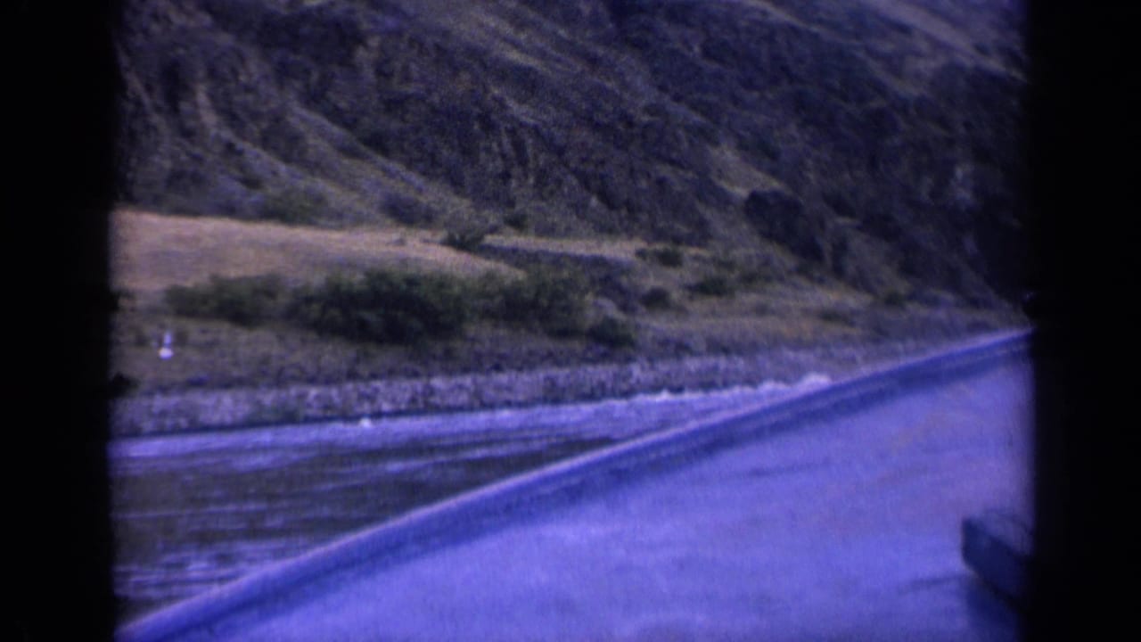 River and road edge passing along a rocky bank beneath a hillside.