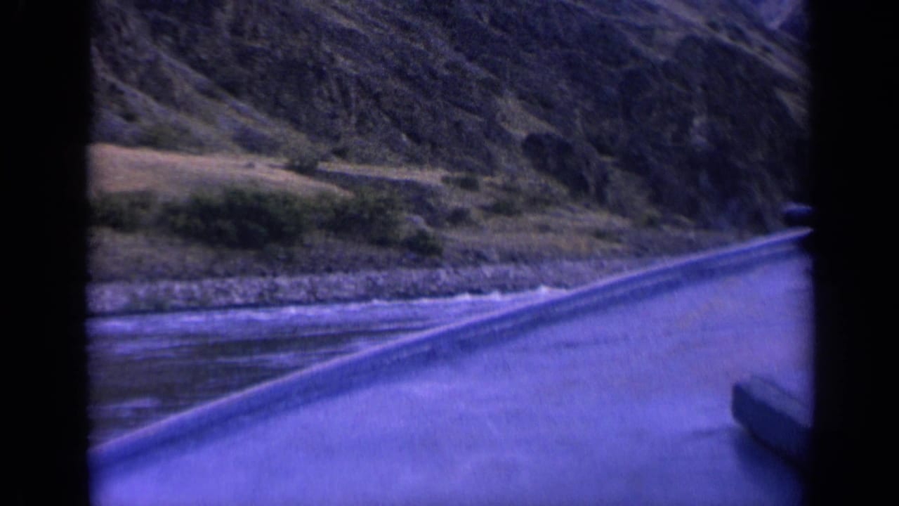 A 1963 Washington home-movie frame shows a blurred roadside river view with rocky shore, low brush, and a steep natural slope.