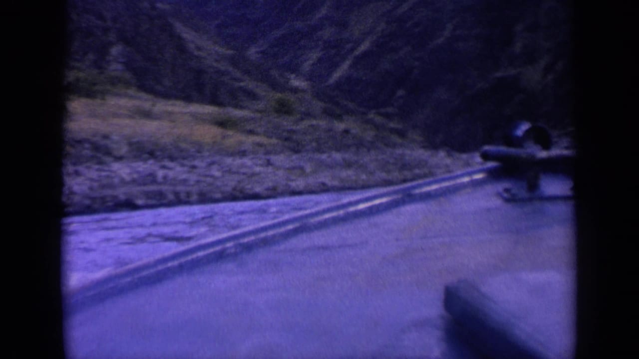 1963 Washington 8mm frame showing the boat-side perspective, trailing wake, rocky bank, and steep mountain slope in blue-purple color cast.